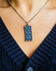 The Women's City Club necklace is a rectangular metal pendant with a geometric design that includes teardrop shapes and smoothly curved lines in various shades of blue. The pendant hangs from a silver chain around the neck of a woman in a navy cable knit sweater.