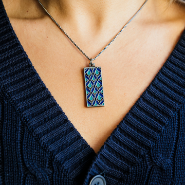 The Women's City Club necklace is a rectangular metal pendant with a geometric design that includes teardrop shapes and smoothly curved lines in various shades of blue. The pendant hangs from a silver chain around the neck of a woman in a navy cable knit sweater.